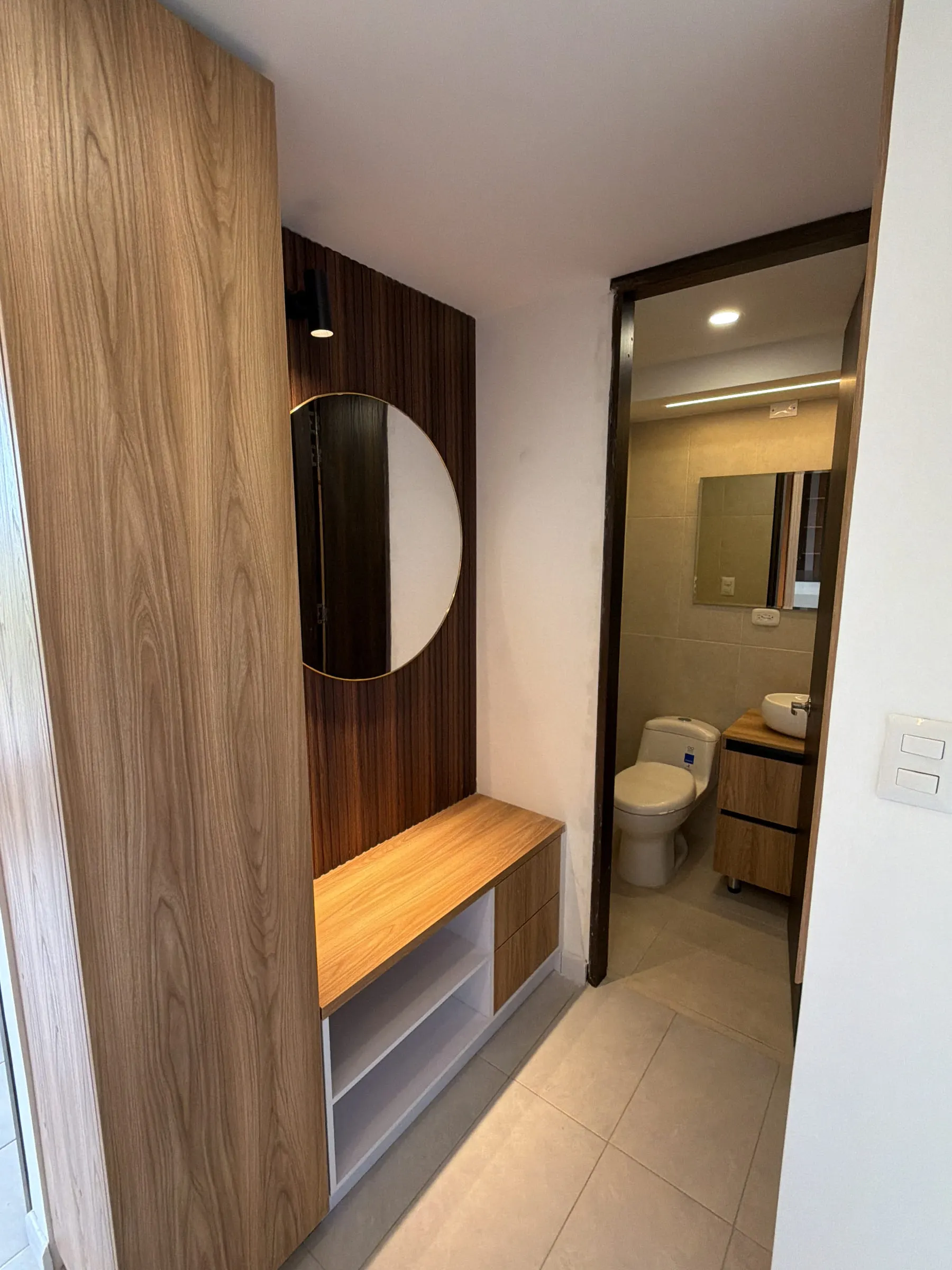 Mueble recibidor en madera con espejo redondo y banca con cajones, junto al acceso del baño.