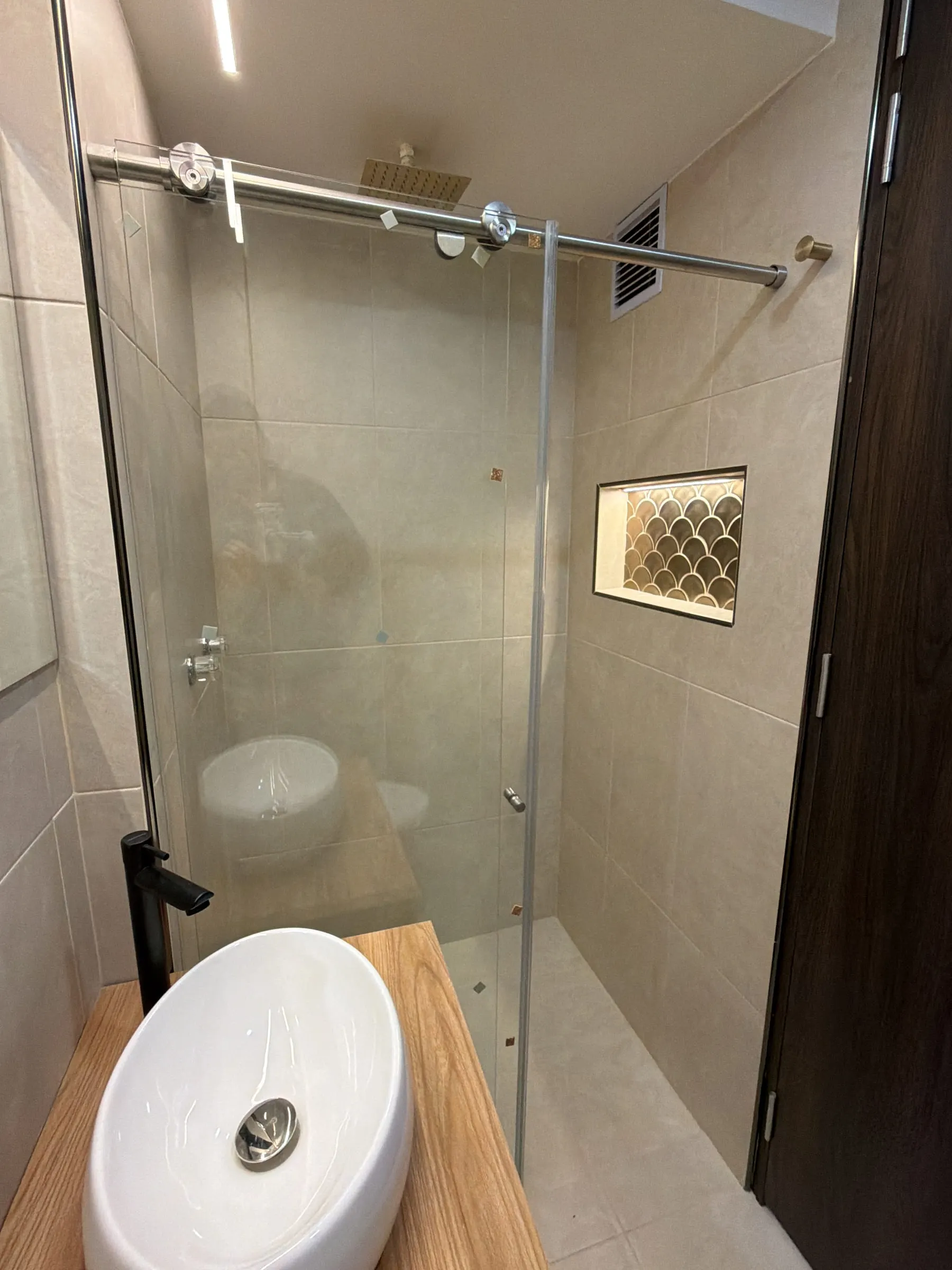 Baño con lavamanos sobrepuesto sobre madera, ducha en vidrio templado y nicho enchapado.
